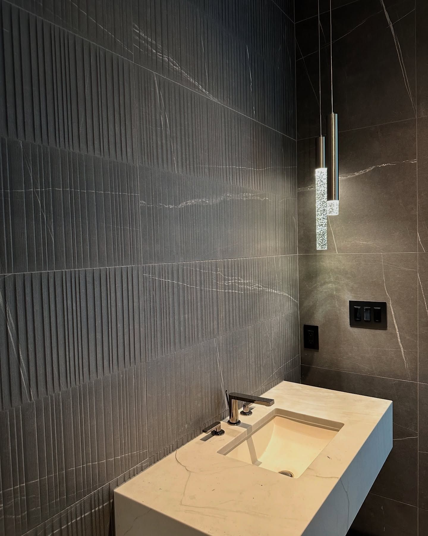Modern bathroom with textured gray wall tiles, light countertop, and pendant lights.