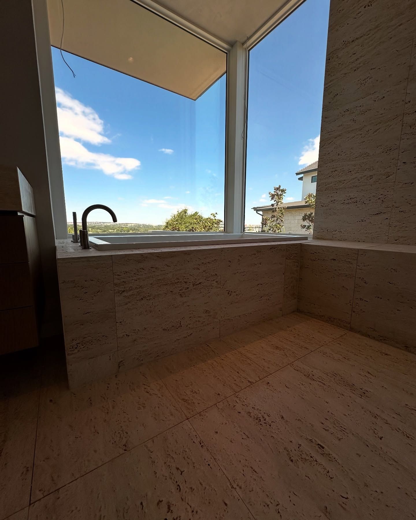 Bathroom with travertine walls and floor, a large window with a view of the sky, and a faucet.