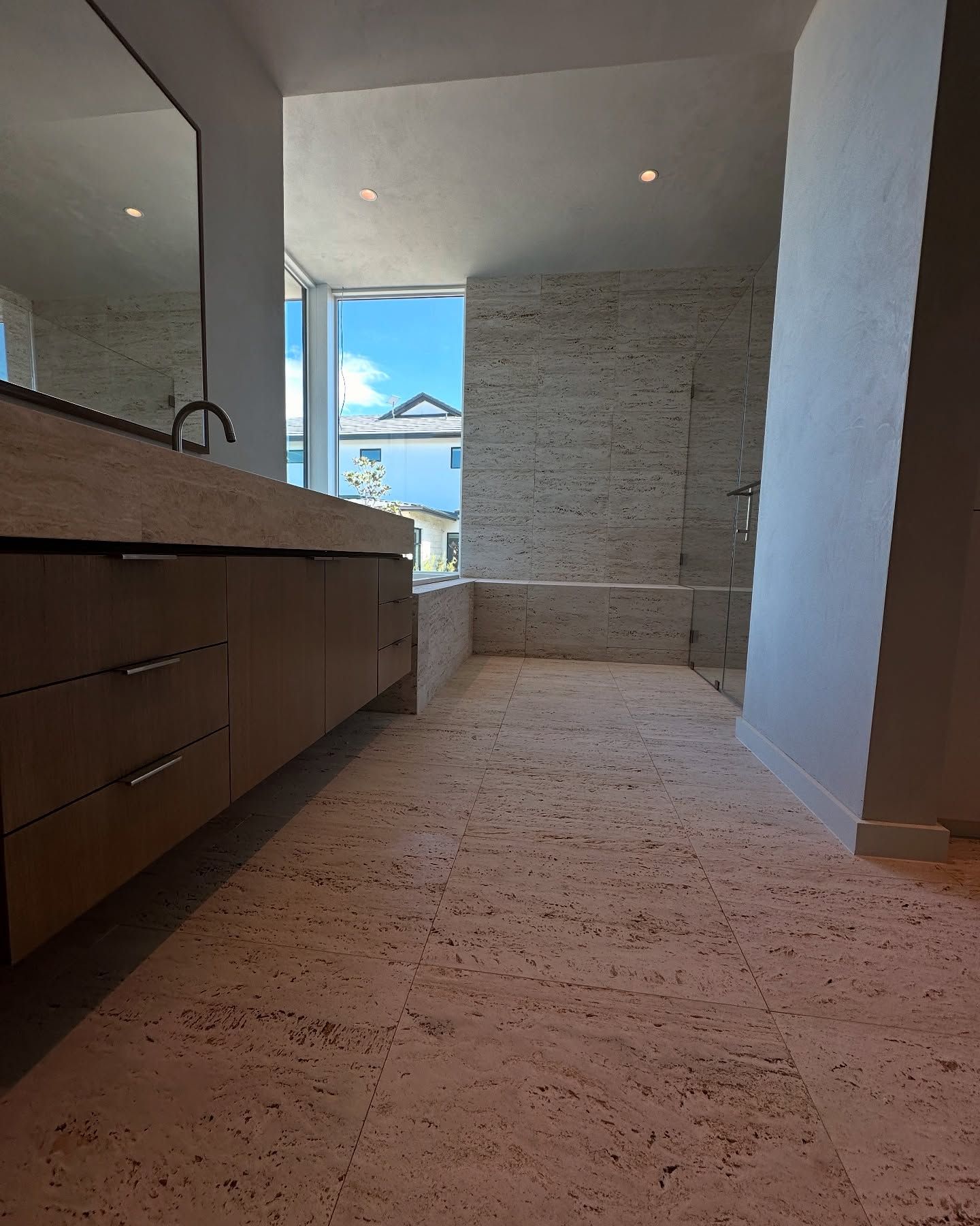 Bathroom with beige stone floor, vanity, and window overlooking a blue sky.