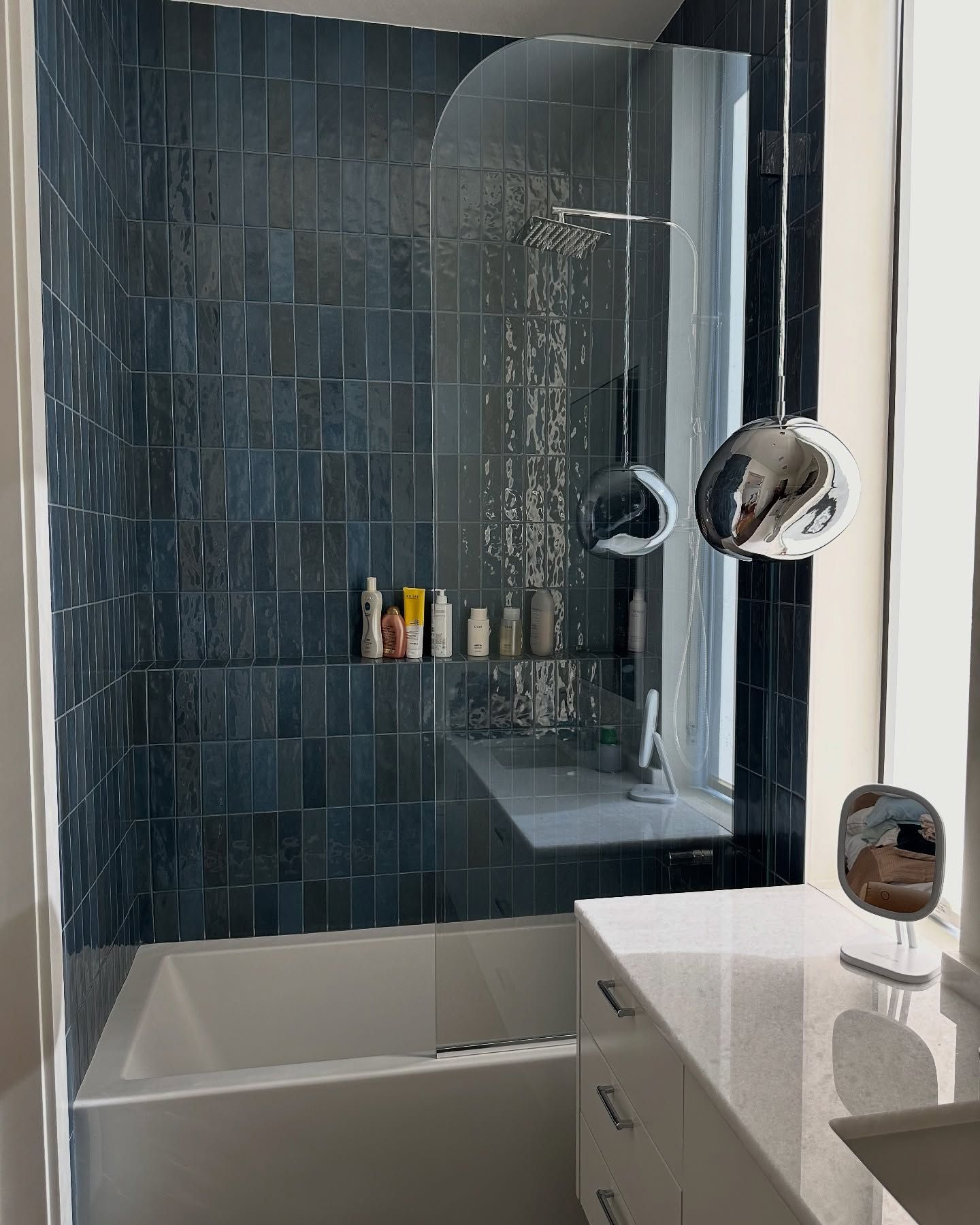 Bathroom with blue tiled shower, glass screen, white tub, and vanity with silver pendant lights.