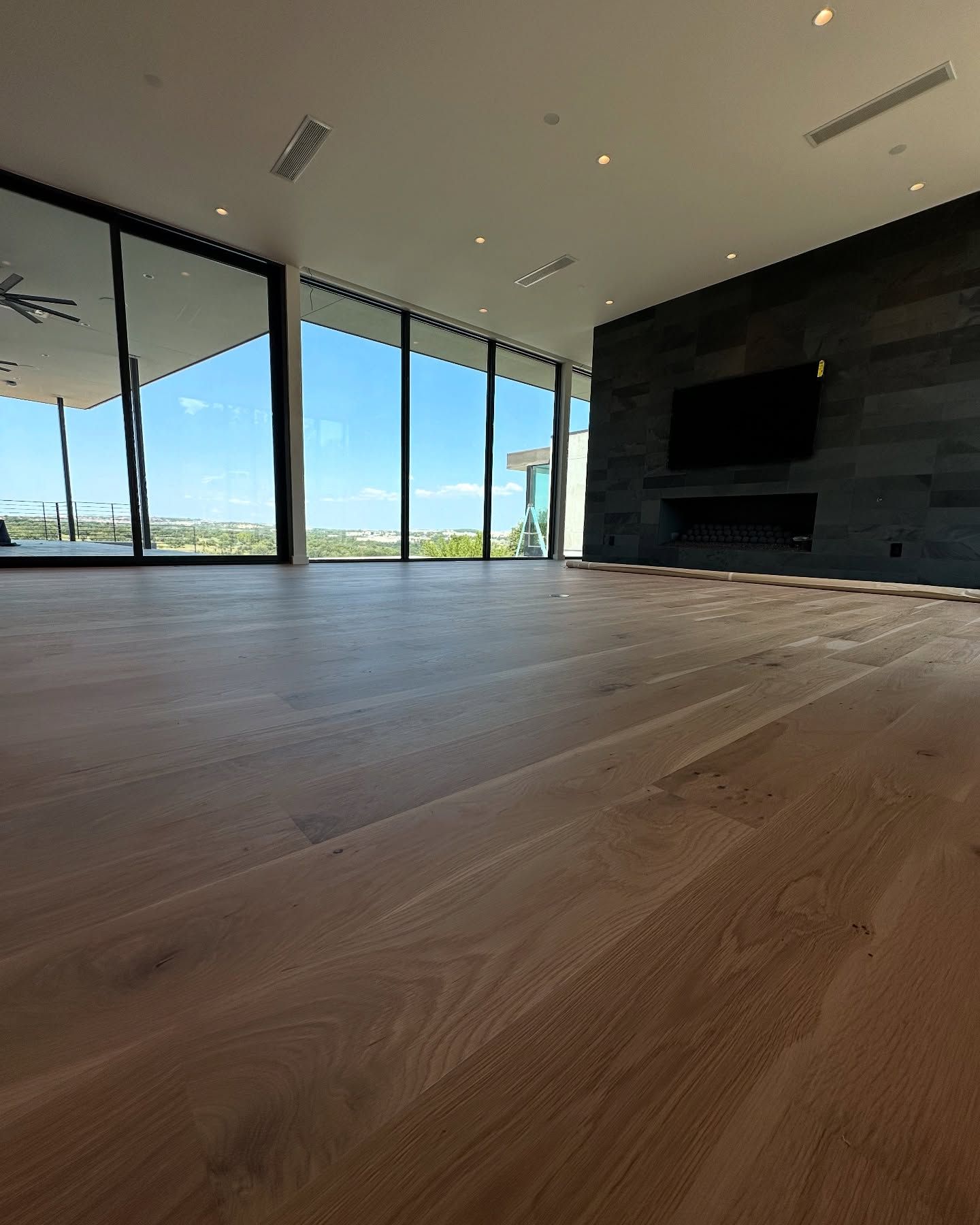 Empty room with wood floors, large windows with a view, and a dark stone fireplace with a TV.
