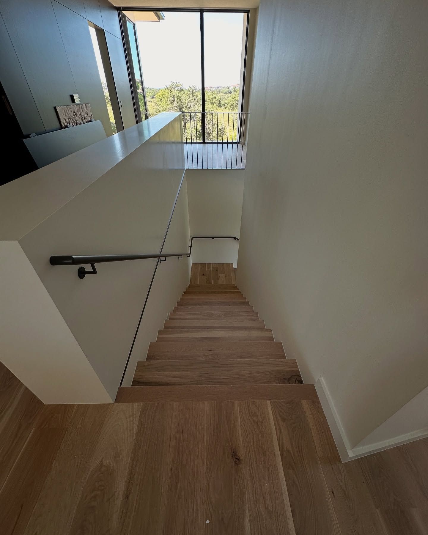Wooden stairs leading down with white walls and a black railing. Large window at the top.