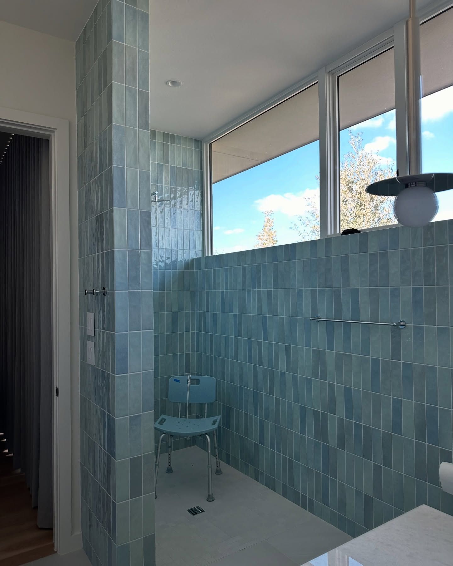Blue-tiled accessible shower with a seat, stainless steel bars, and large window.