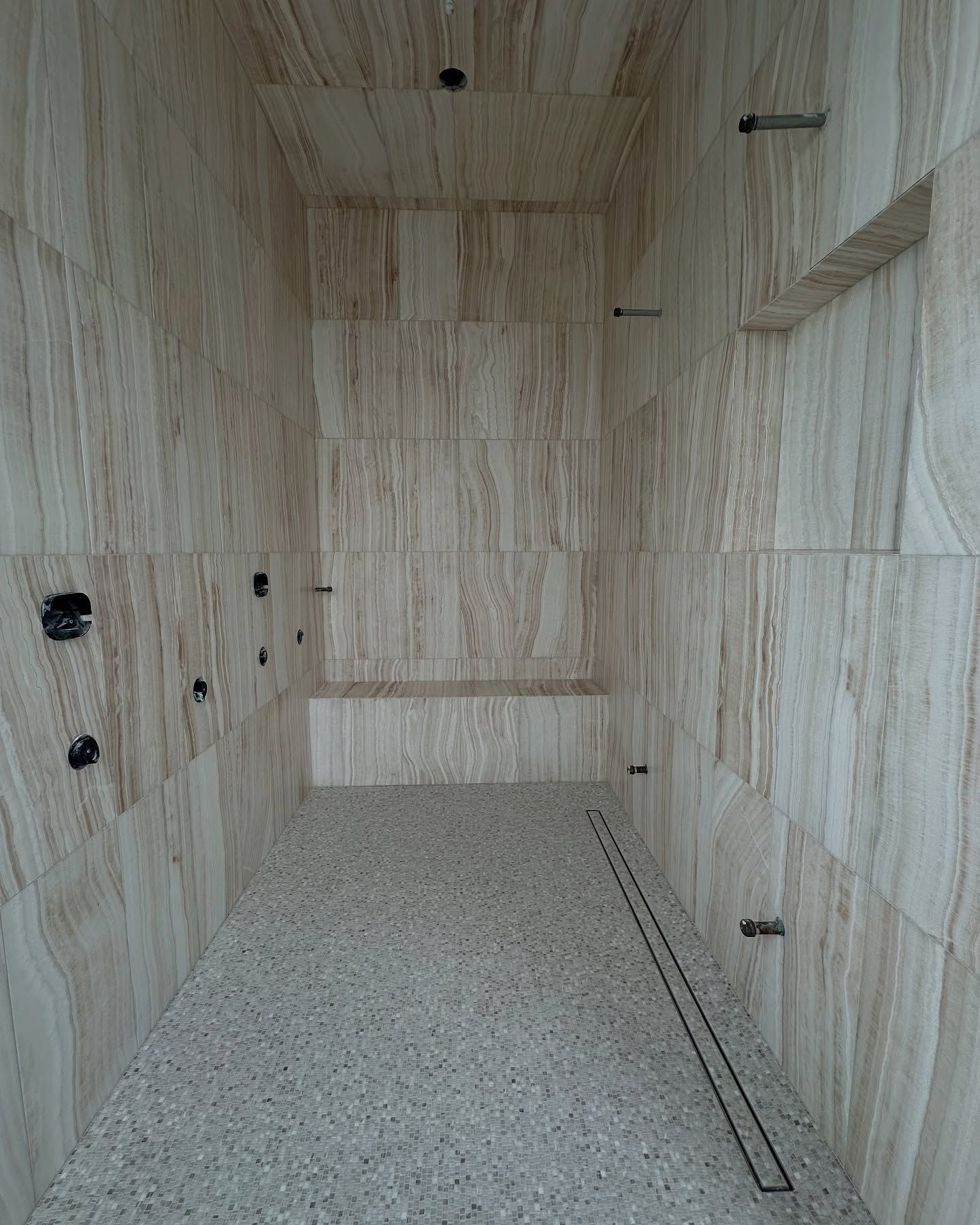 An empty shower with beige marble walls, gray speckled floor, and a linear drain.