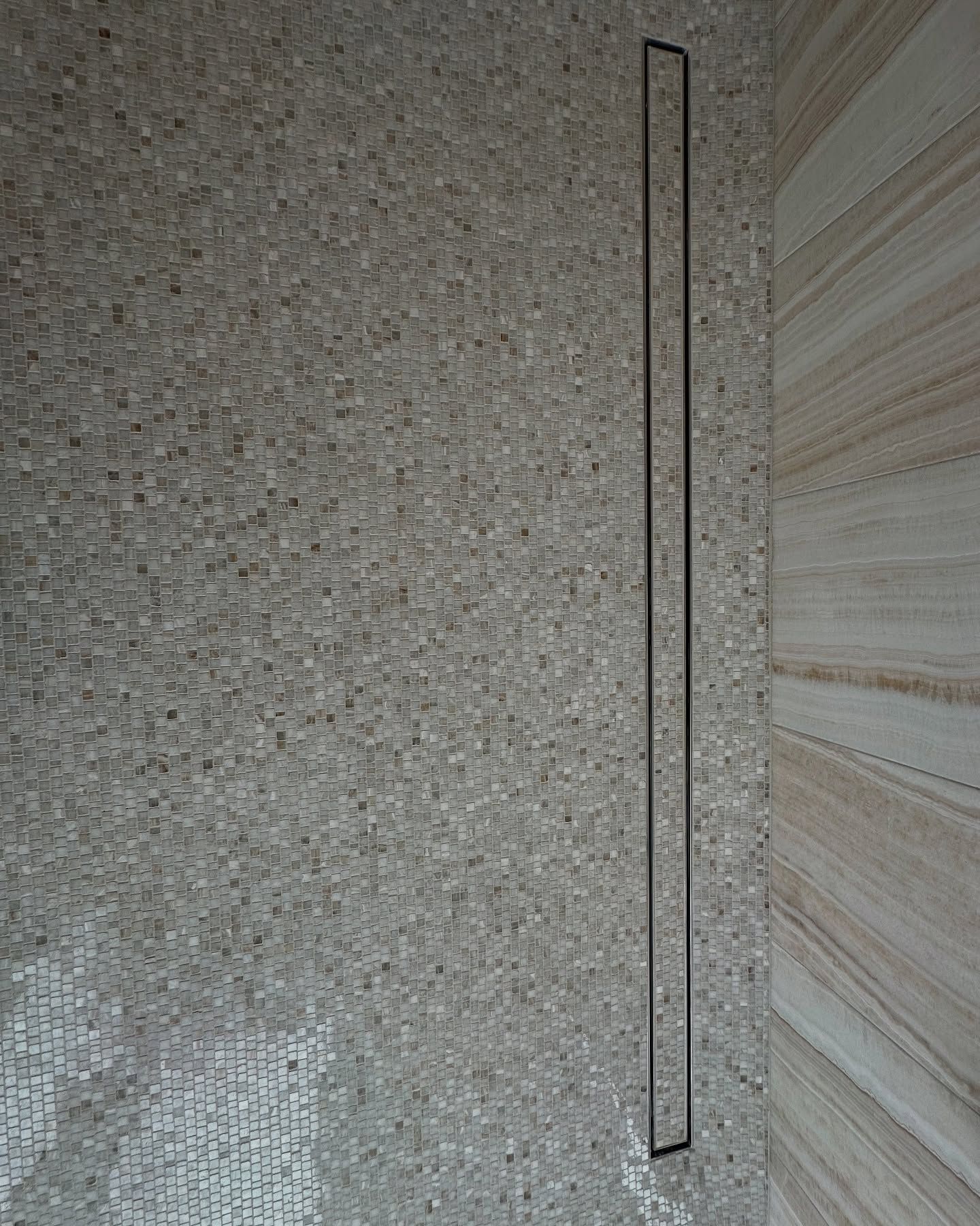 Long, thin, black shower drain on a wall with beige and brown speckled tile.