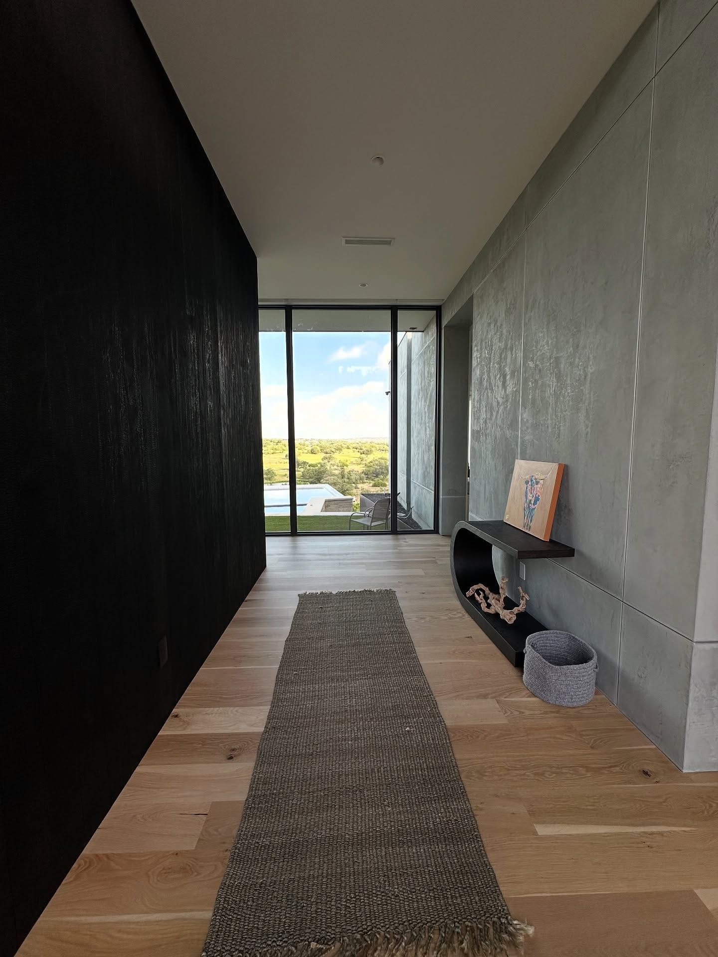 Hallway with wood floor, textured walls, rug, table, and large window overlooking trees.