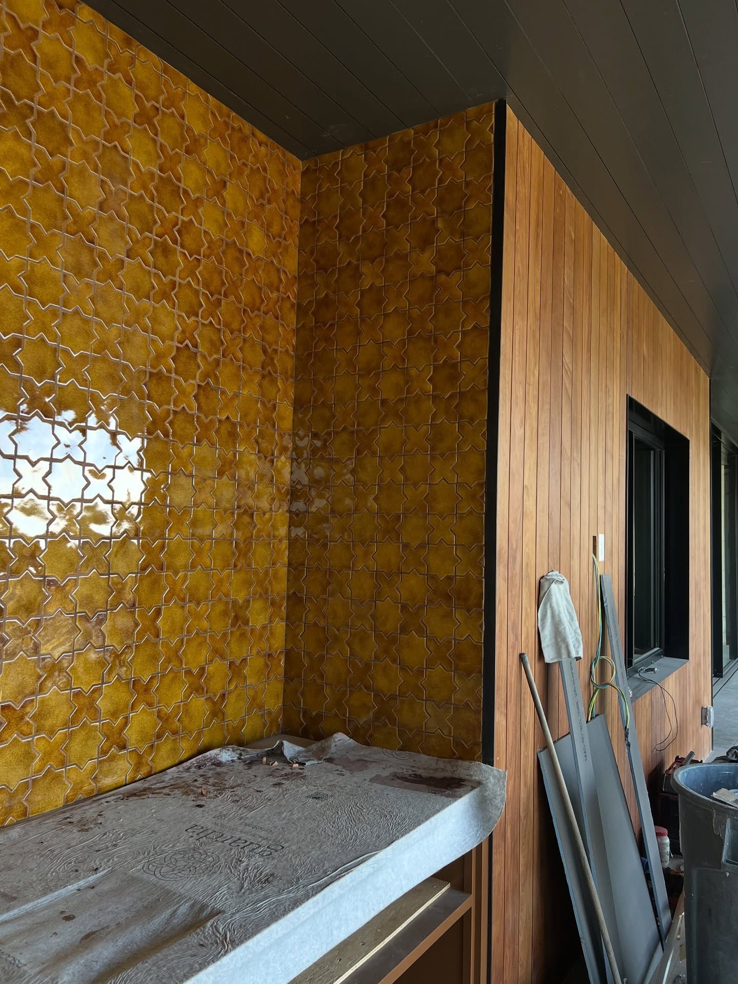 Golden mosaic tile wall and wooden paneling in a corner with a partially visible window and construction materials.