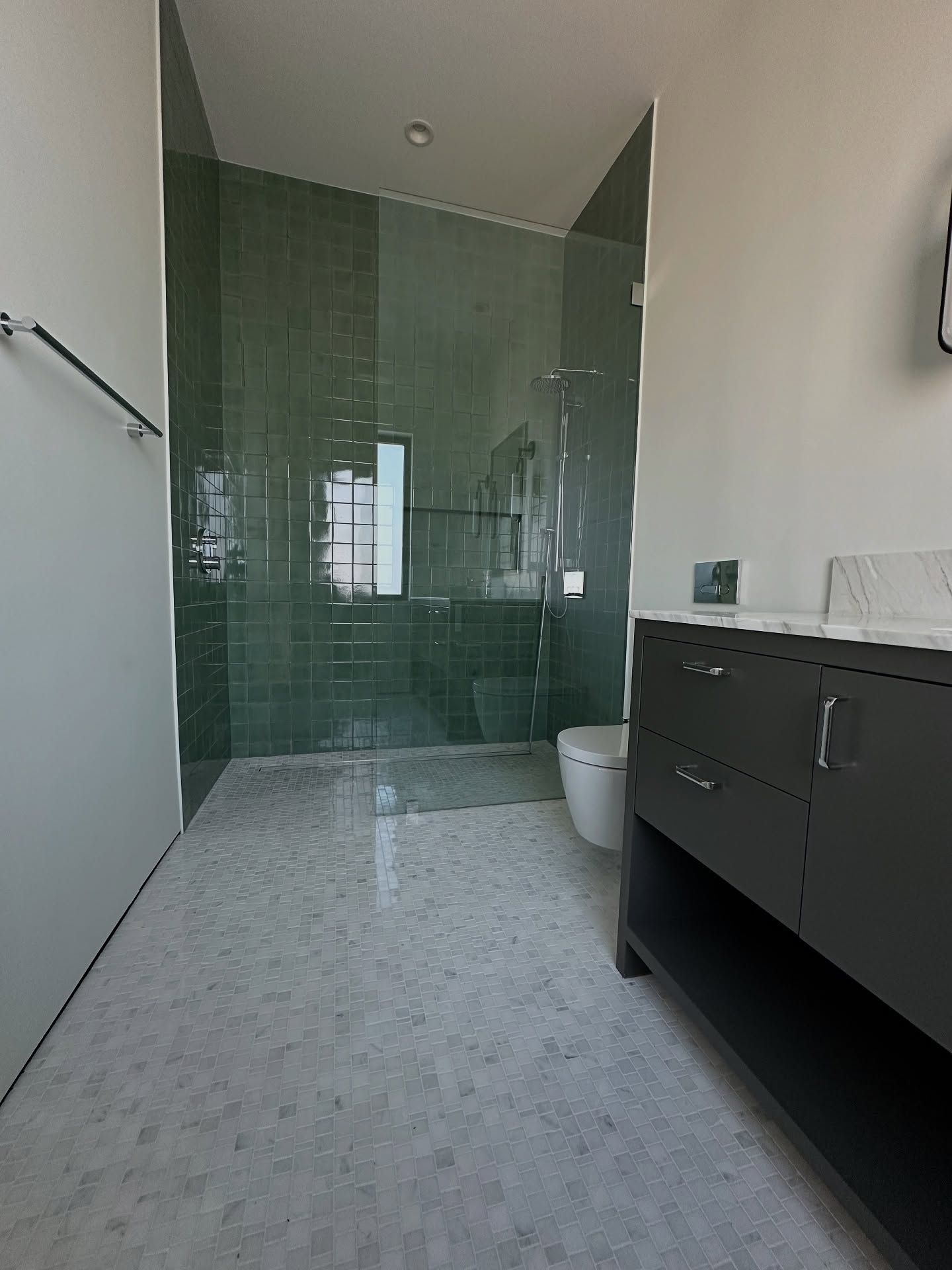 Green-tiled shower with glass door and a dark vanity in a bathroom with terrazzo flooring.