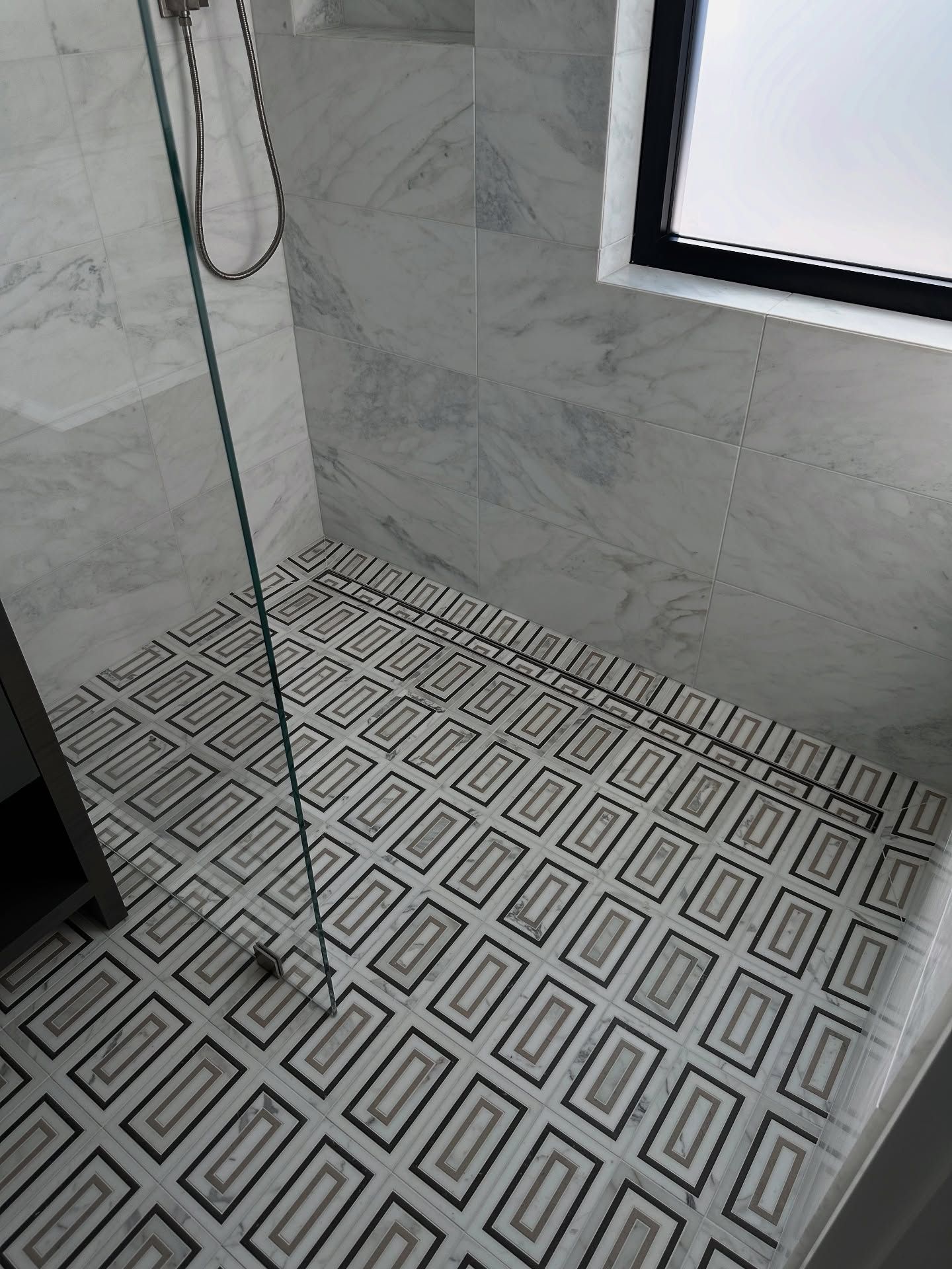 Tiled shower with a patterned floor and glass door. White and gray marble-look walls, and a window in the upper right.