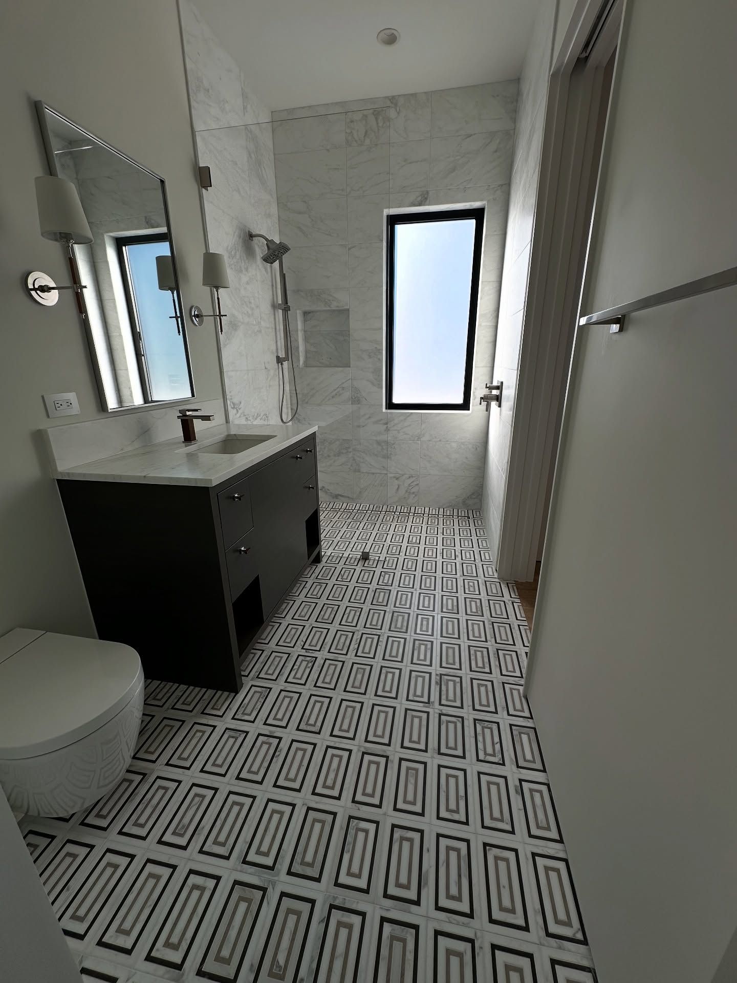 Bathroom with black and white patterned floor tiles, marble shower, dark vanity, and window.