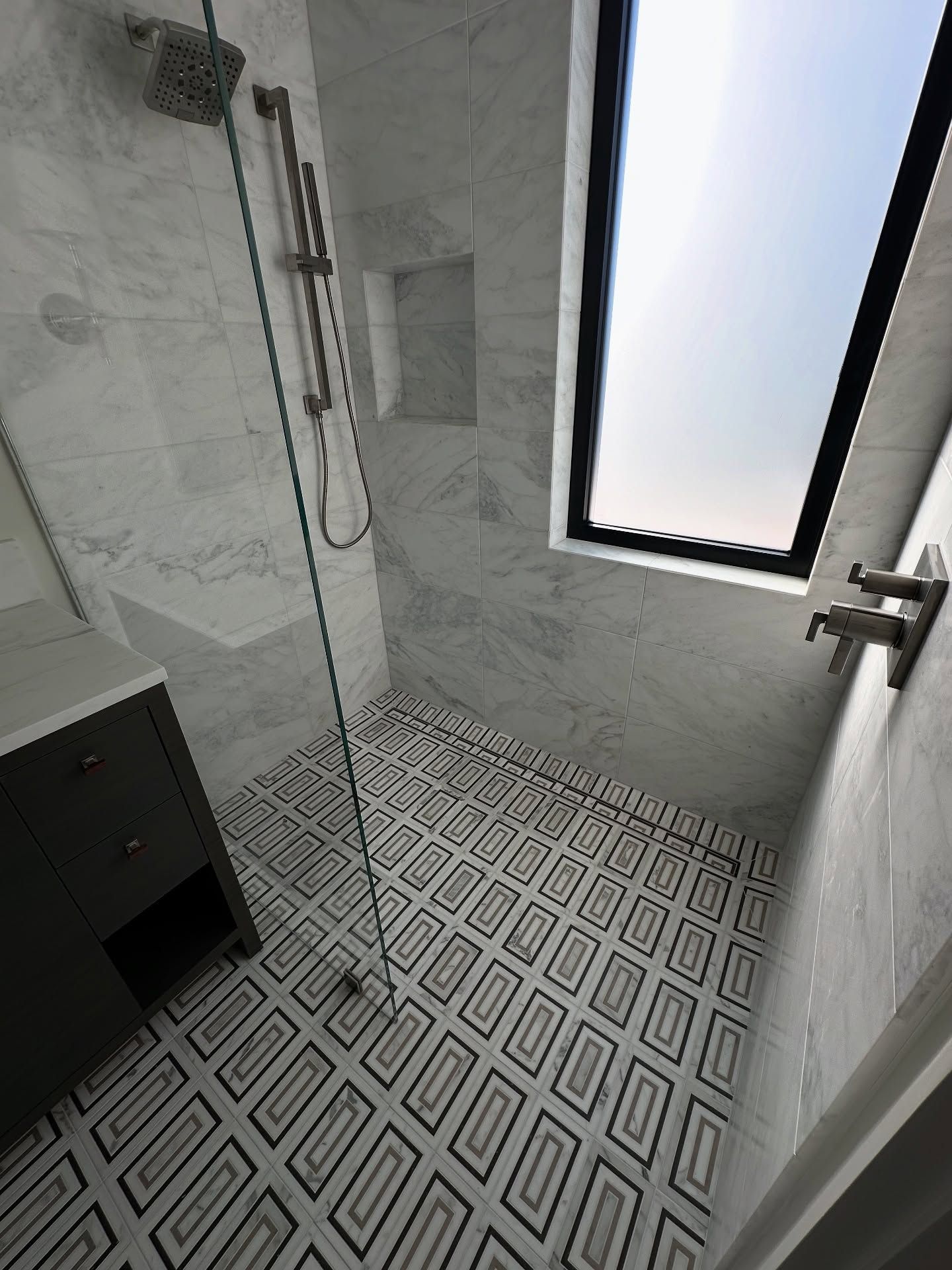 Modern bathroom with patterned tile floor, marble shower walls, and a skylight. Gray vanity on left.