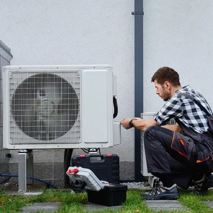 A technician in work coveralls kneeling outdoors to repair a white air conditioning unit next to a toolkit.