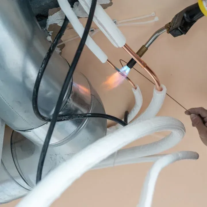 A person brazes copper pipes near a metal ventilation duct, heating the joint with a propane torch.