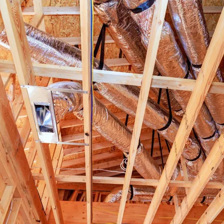 Flexible HVAC ductwork and a metal register box installed between wooden ceiling joists in an unfinished home.