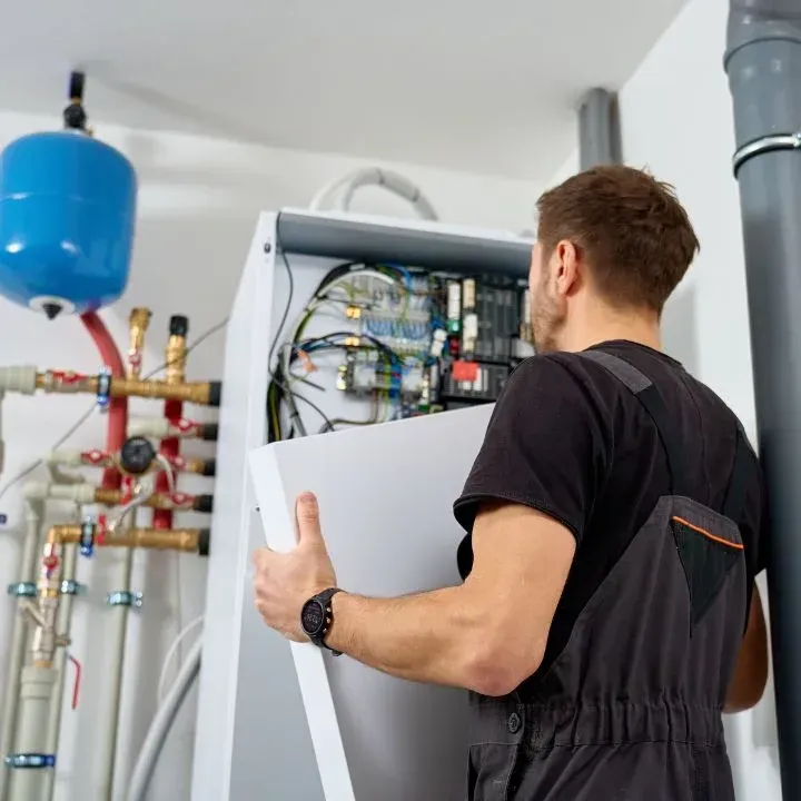 A technician in dark work clothes attaches a cover to an open HVAC unit, with plumbing components visible in the background.