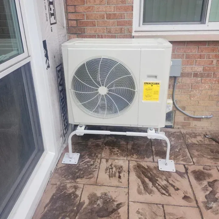 A beige outdoor HVAC unit mounted on a white metal stand on a paved patio against a brick wall.