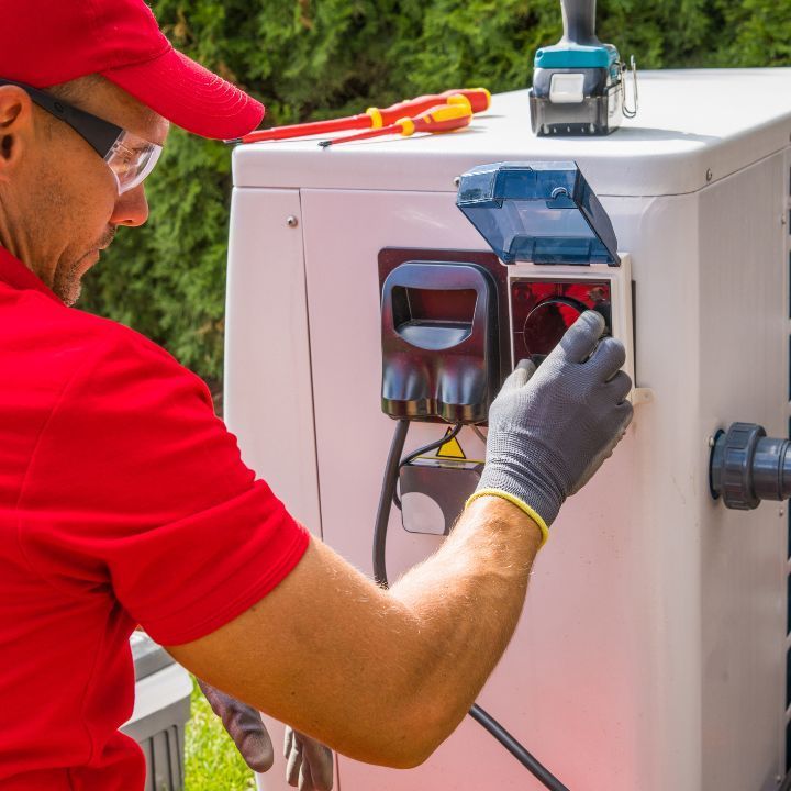 A technician in a red uniform and protective gloves services an outdoor heat pump unit with professional tools.