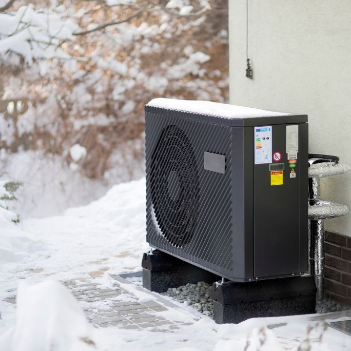 A grey air-source heat pump sits on black mounting blocks outside against a wall during a snowy winter day.