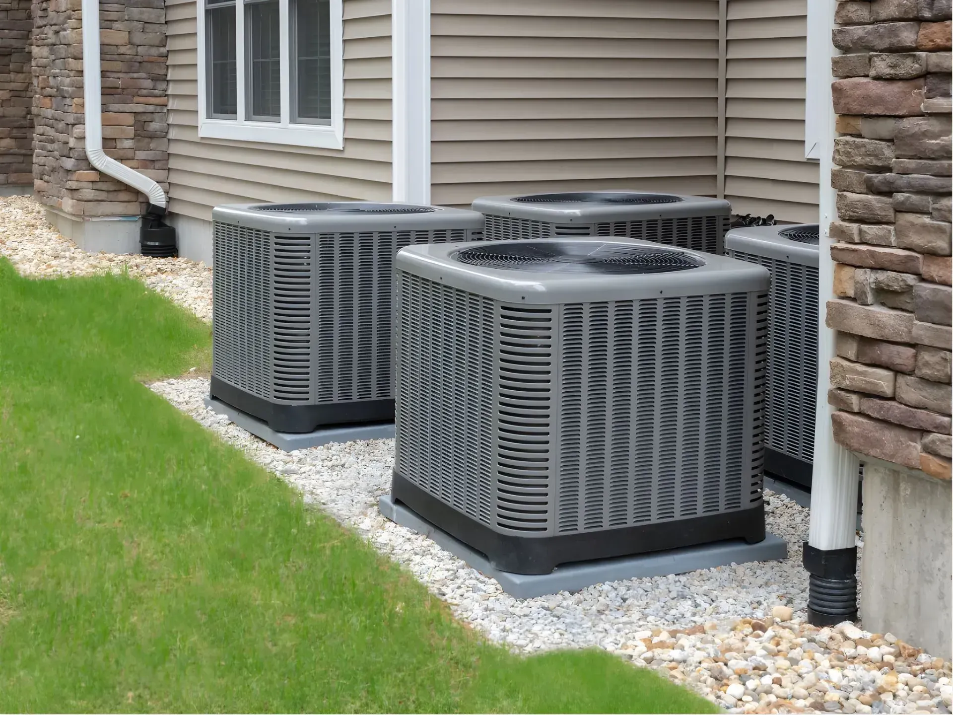 Four central air conditioning units near a building's exterior, surrounded by grass and small stones.