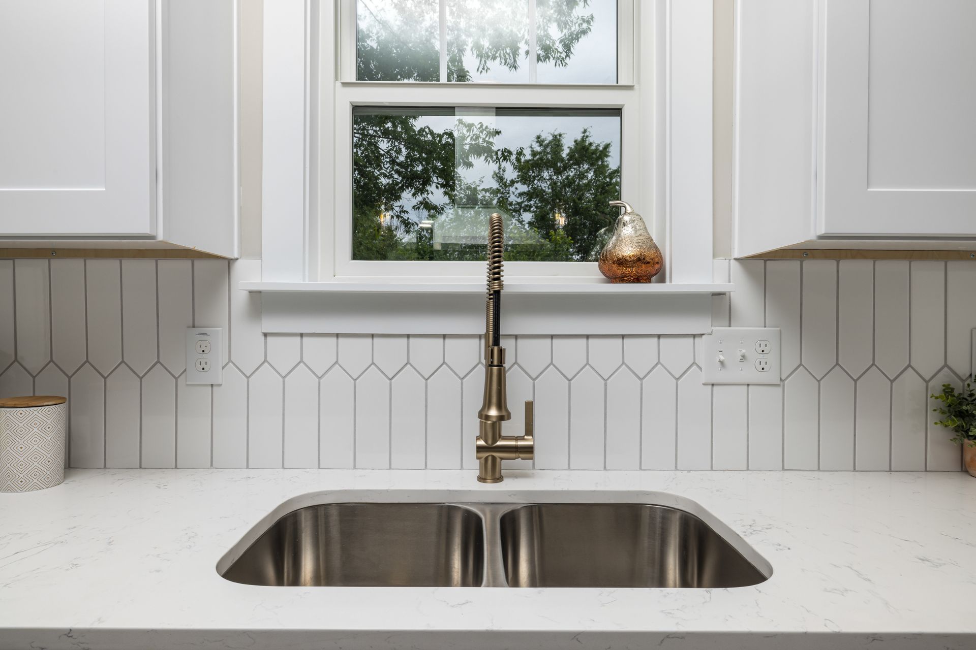 Kitchen sink with stainless steel basin, gold faucet, white cabinets, and backsplash with window. Plumbing services in Nothville, MI