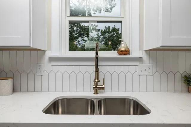 Kitchen sink with stainless steel basin, gold faucet, white cabinets, and backsplash with window. Plumbing services in Nothville, MI