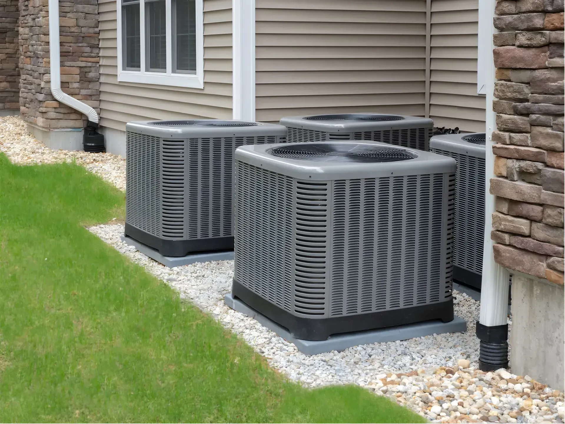 Two outdoor air conditioner units beside a beige house with stone siding and green lawn