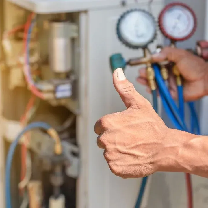 A technician gives a thumbs up while holding a manifold gauge set, indicating repair of an HVAC unit's outdoor compressor.