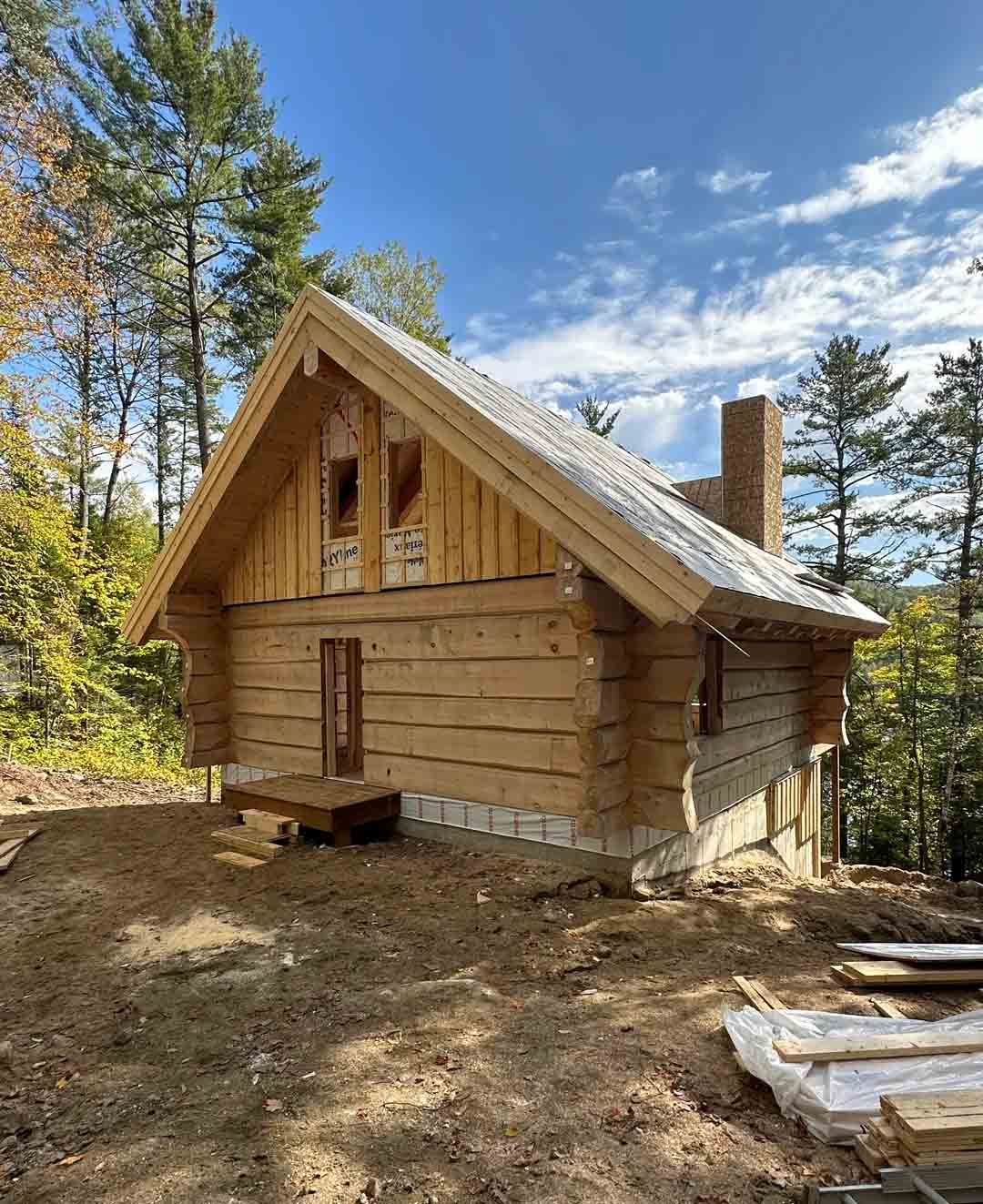Petite cabane en rondins en construction dans une zone boisée, avec un toit en métal et une cheminée.