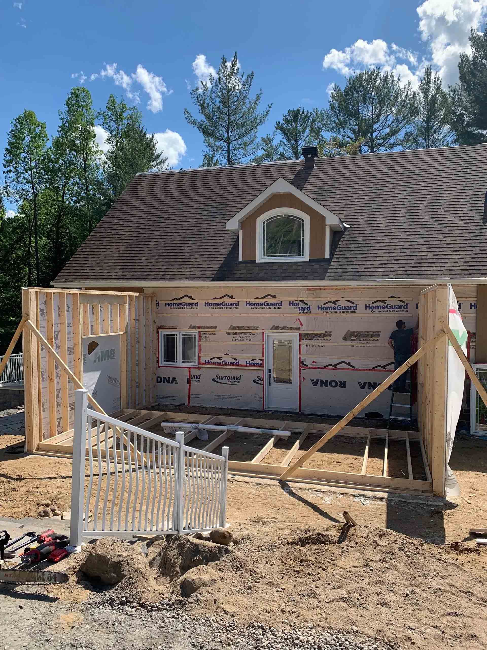 Maison en construction avec charpente apparente, bardage blanc et toit à lucarnes sous un ciel bleu.