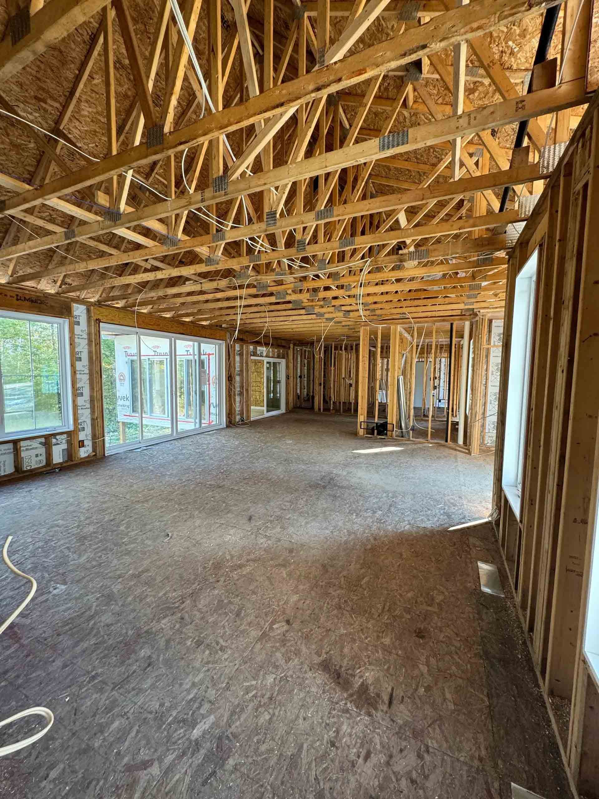 Intérieur d'une maison inachevée avec charpente en bois apparente, fermes et grandes fenêtres sur un côté.