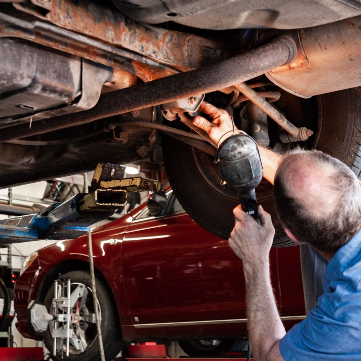 Mechanic working under a red car, using a power tool to inspect vehicle components.