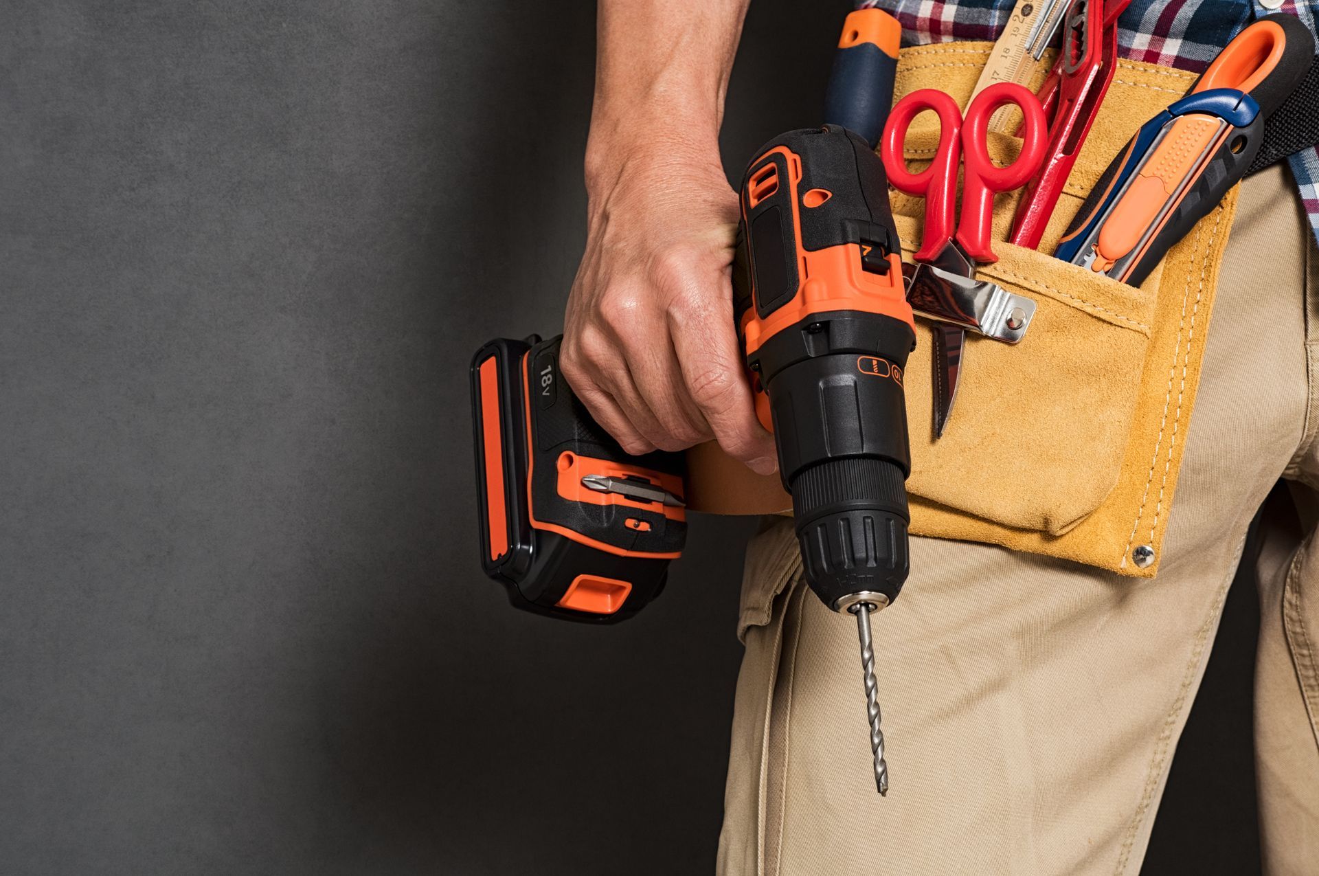 Person holding a cordless drill with tool belt of pliers and scissors against a dark background