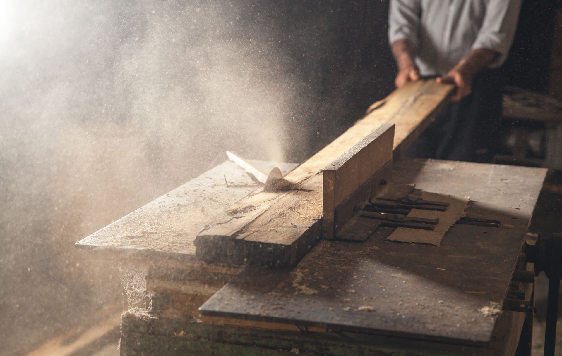Woodworker cutting a plank with a hand saw, sawdust flying in a workshop.