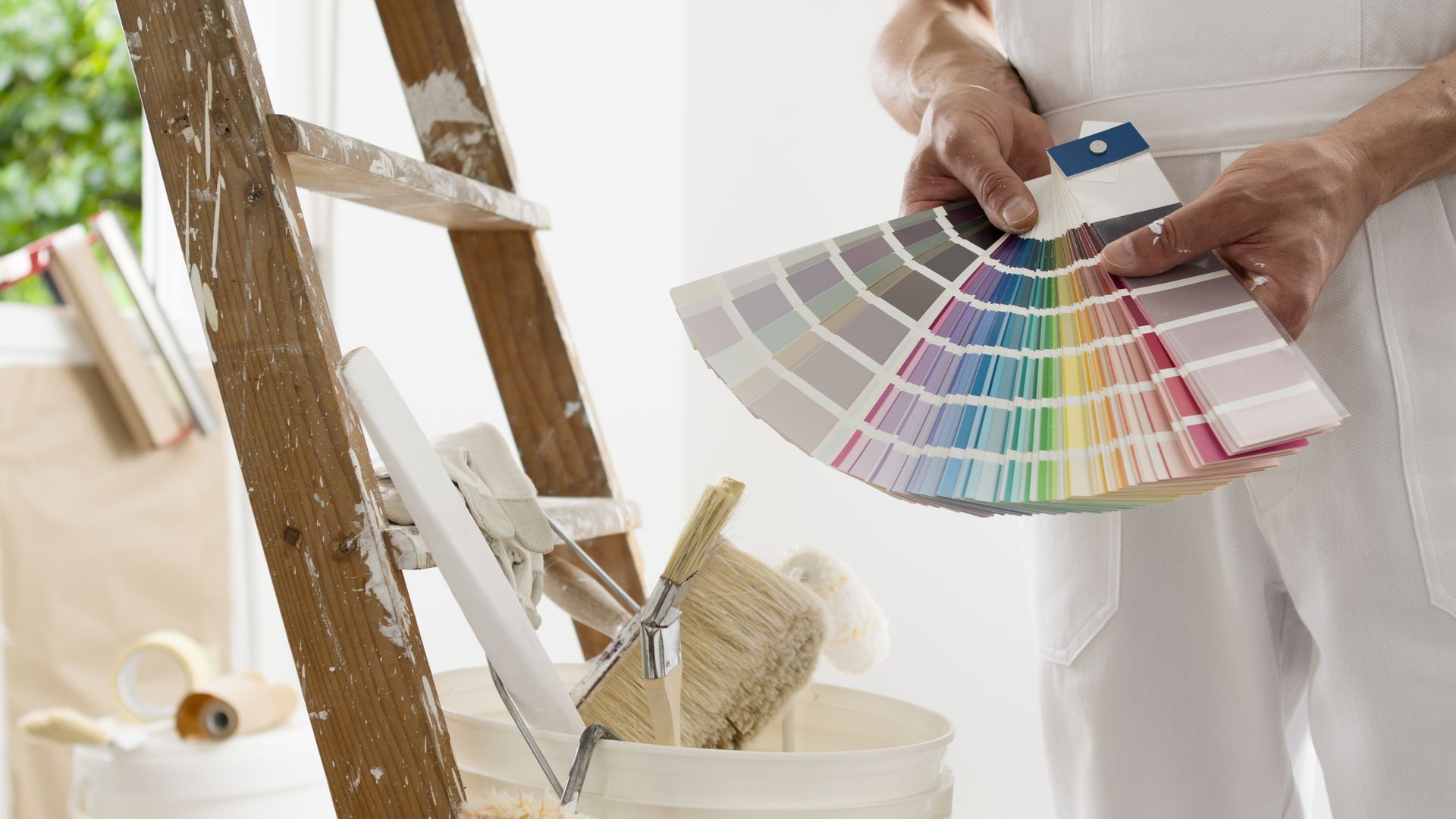 Person holding a fan of paint swatches beside a ladder in a bright room