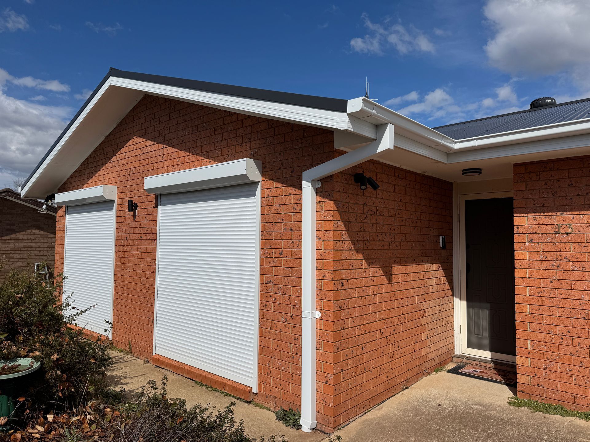 Red brick house with white roller shutters and guttering, blue sky background — All Top Roofing In Tuggeranong, ACT