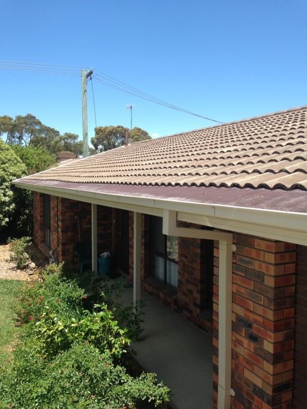 Brick house with brown tiled roof, beige gutters, and green foliage under a clear blue sky — All Top Roofing In Woden, ACT