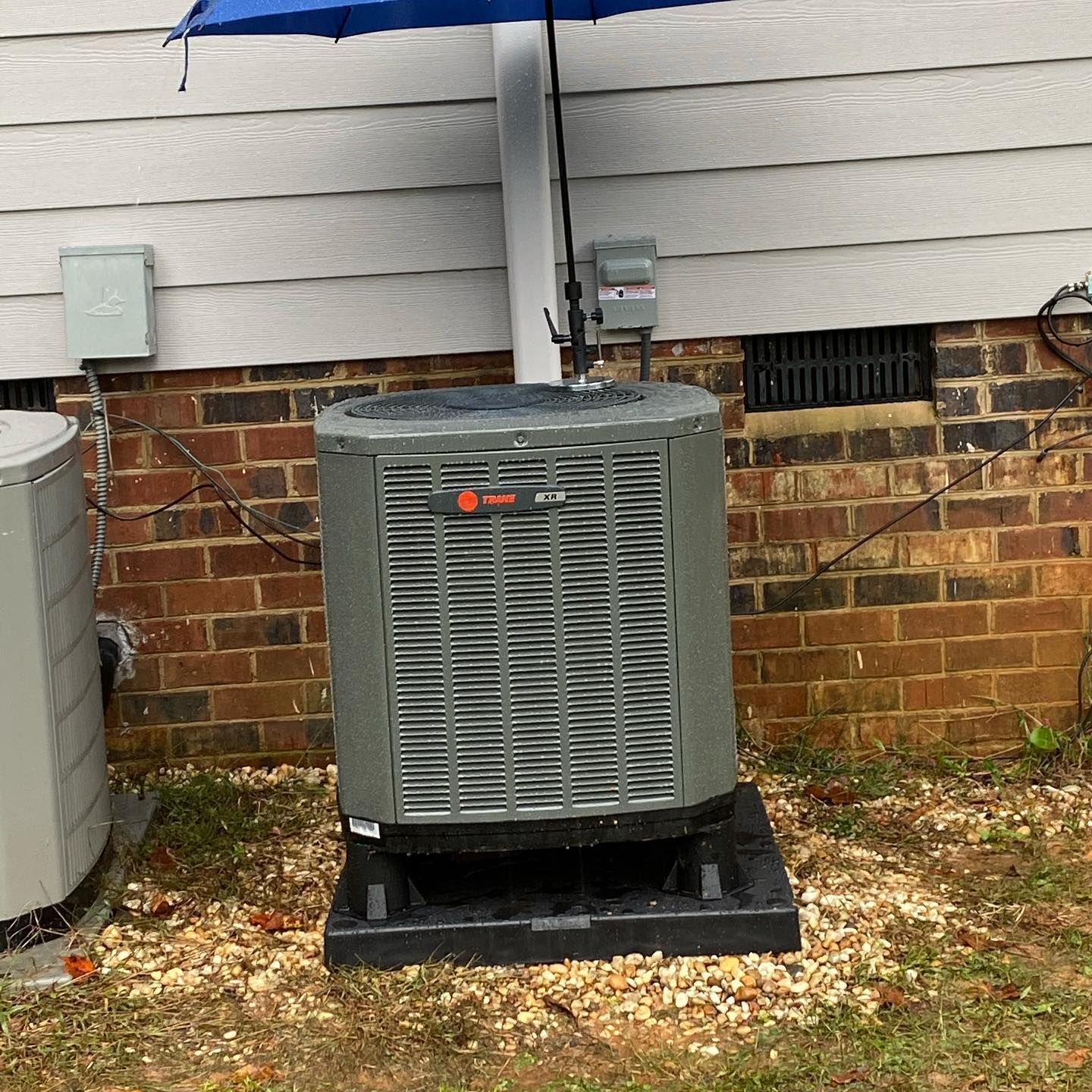 An air conditioner is sitting under an umbrella in front of a brick house.