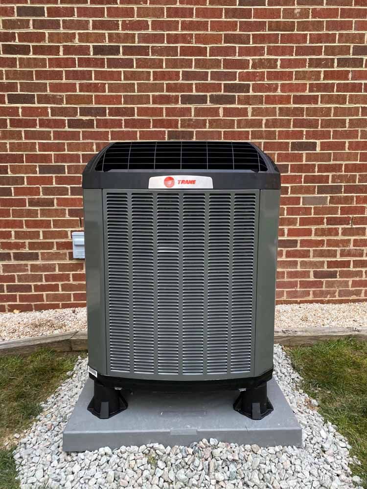 A large air conditioner is sitting on top of a gravel base in front of a brick wall.