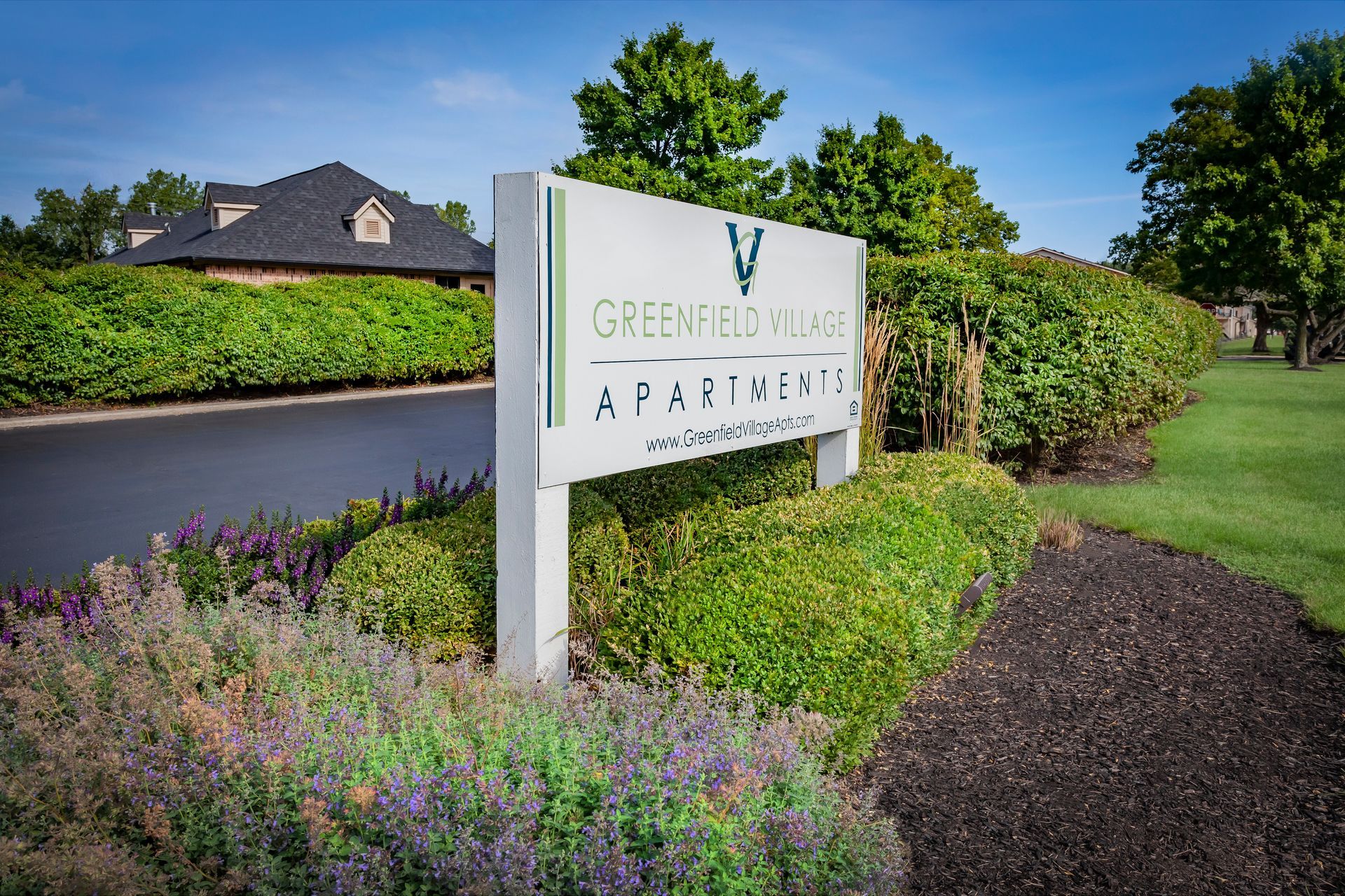 a sign for greenfield village apartments is surrounded by flowers  and bushes at Greenfield Village.
