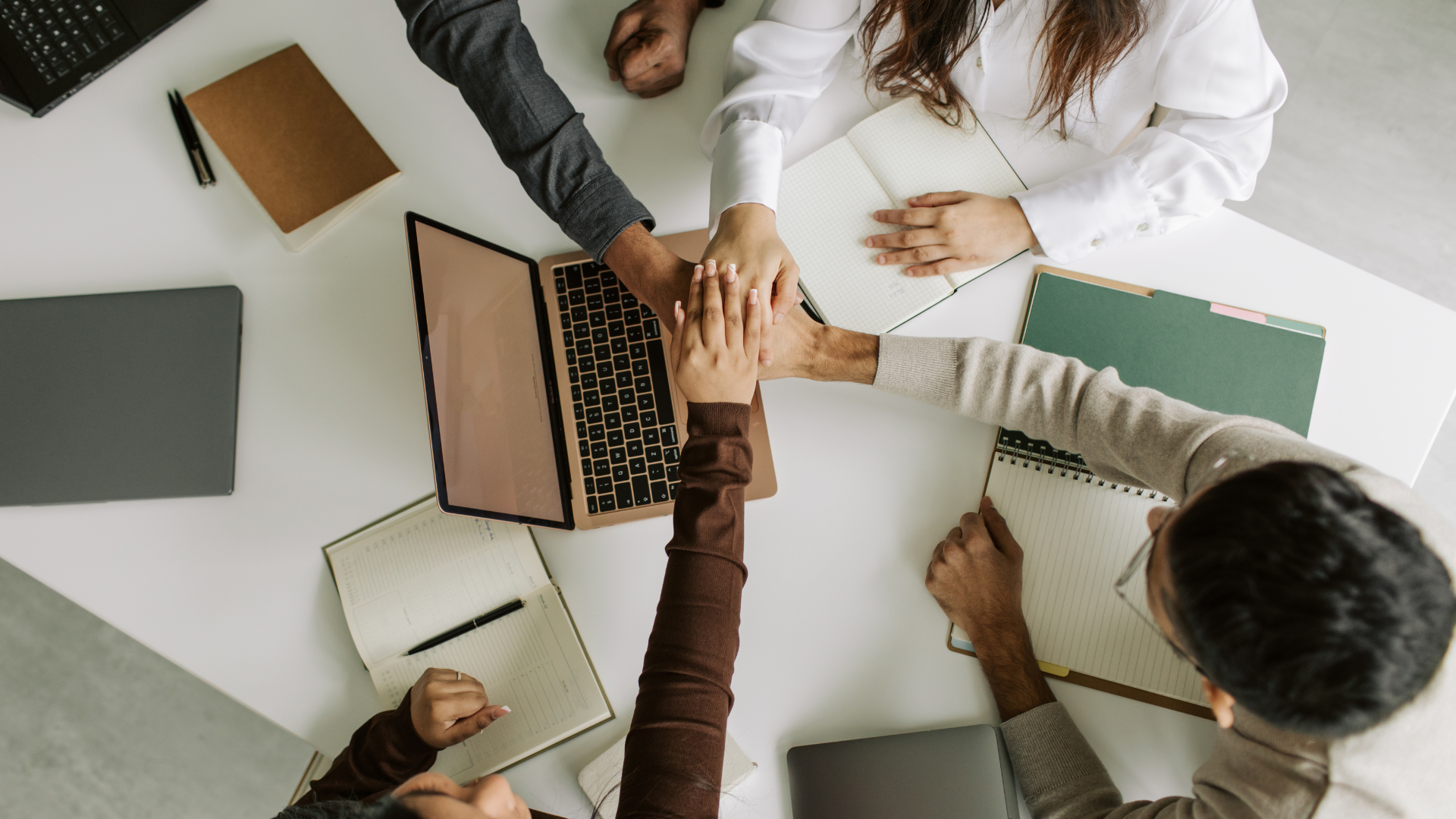 Hands of diverse people stacked together at a table with laptops and notebooks, signifying teamwork.