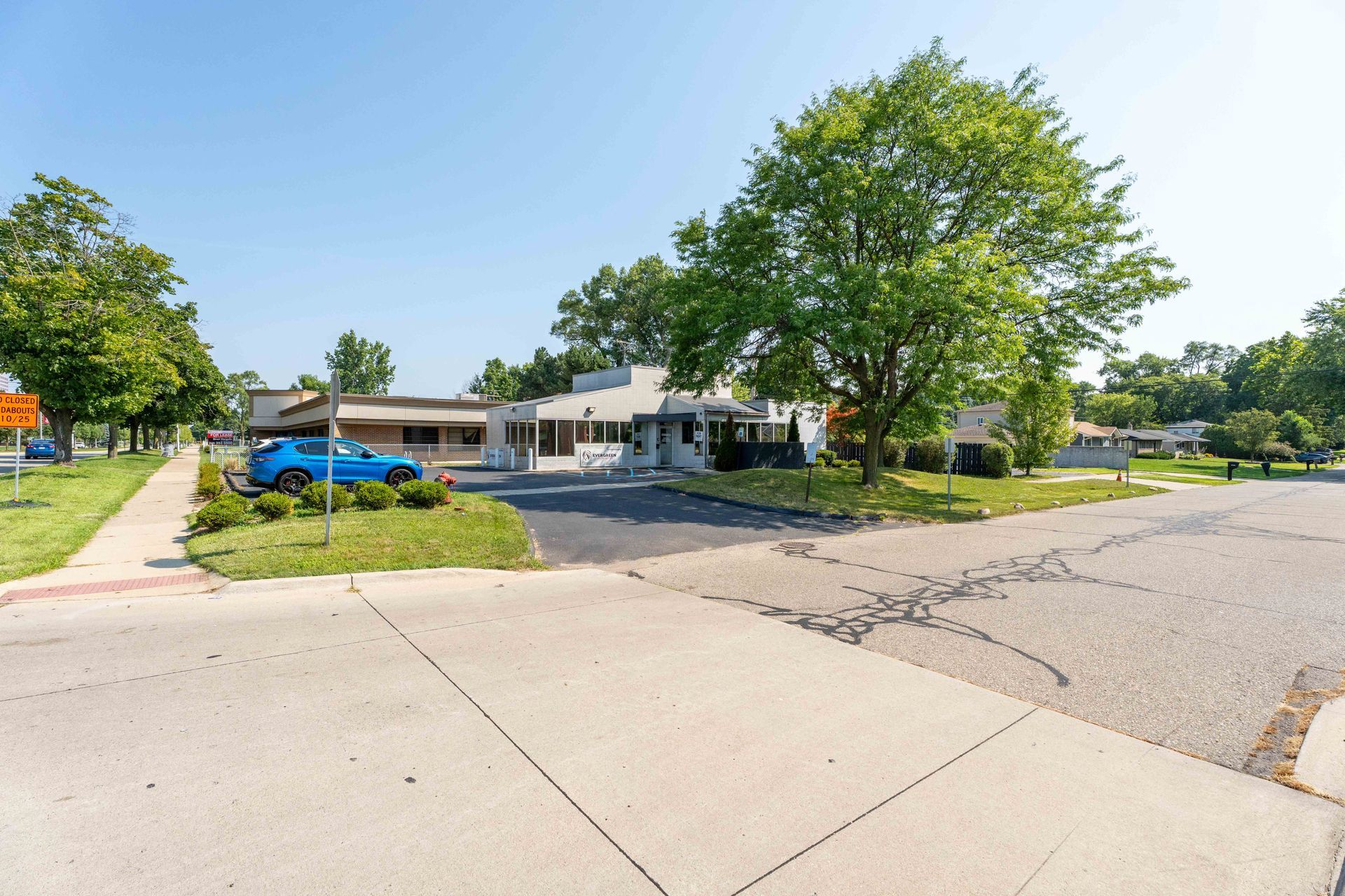 Blue car parked in front of a low, beige building with a small lawn, trees, and sidewalk on a sunny day.