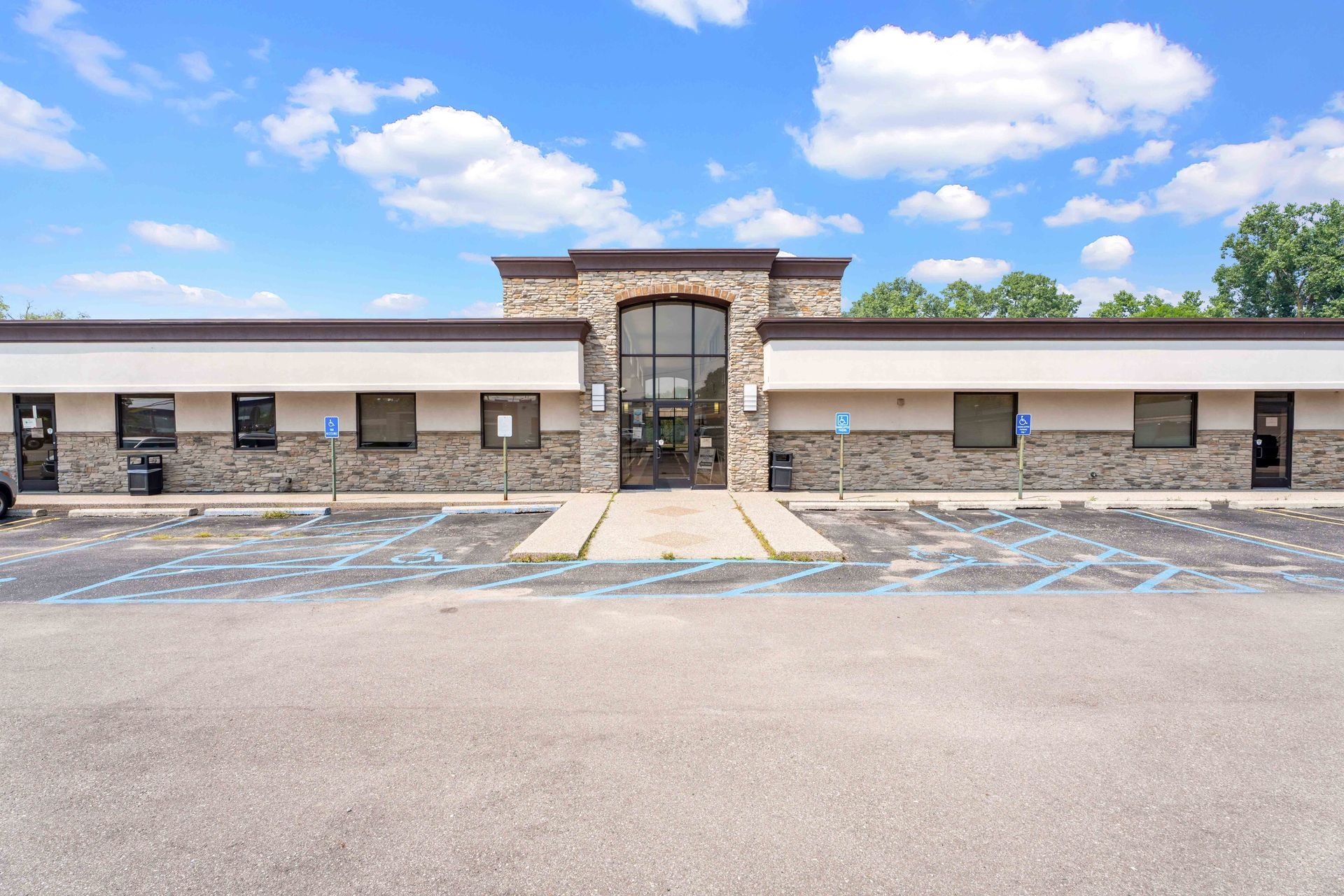 Exterior of a single-story building with a brown stone facade, glass doors, and a paved parking lot.