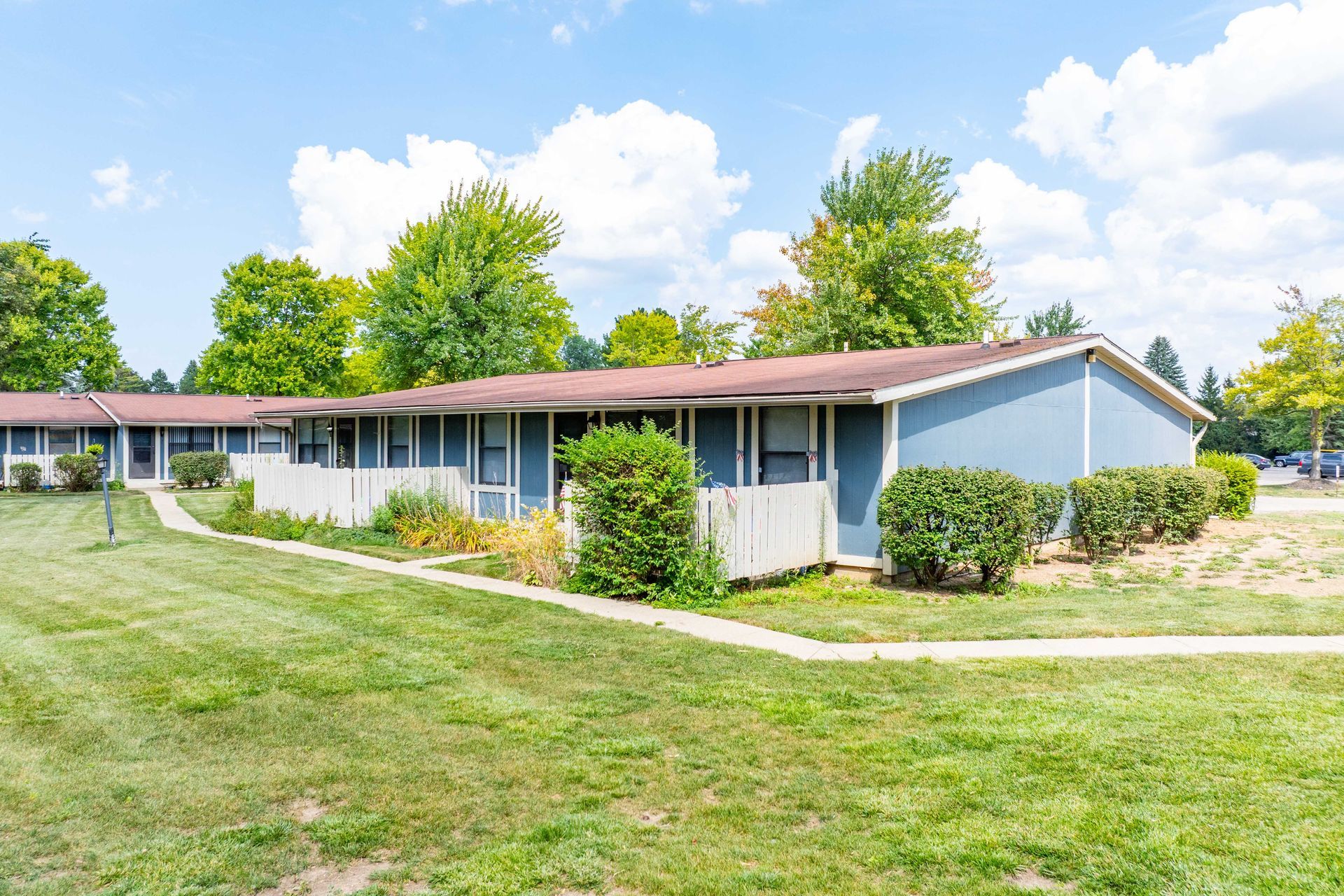 Blue apartment buildings with white fences, surrounded by green grass and trees under a blue sky.