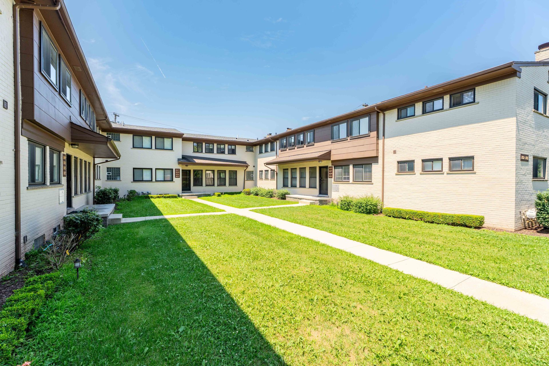 Apartment complex courtyard with green grass, walkways, and light brick buildings under a blue sky.
