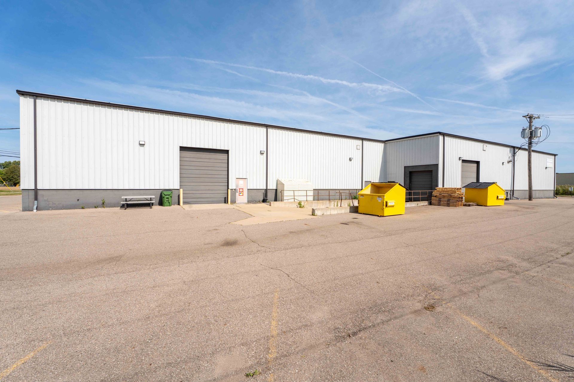 White industrial building with roll-up doors, yellow dumpsters, and a gravel parking lot under a blue sky.