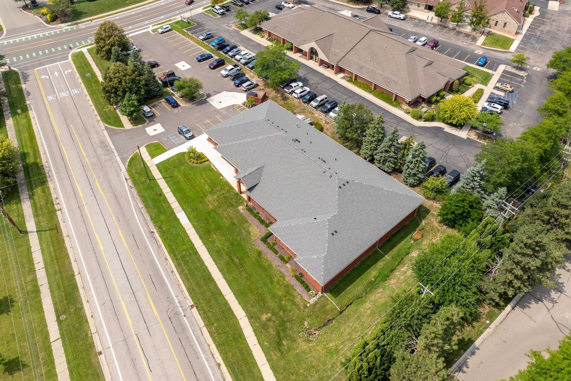 Aerial view of a one-story brick building with a dark roof and green lawn, near a street with cars and trees.