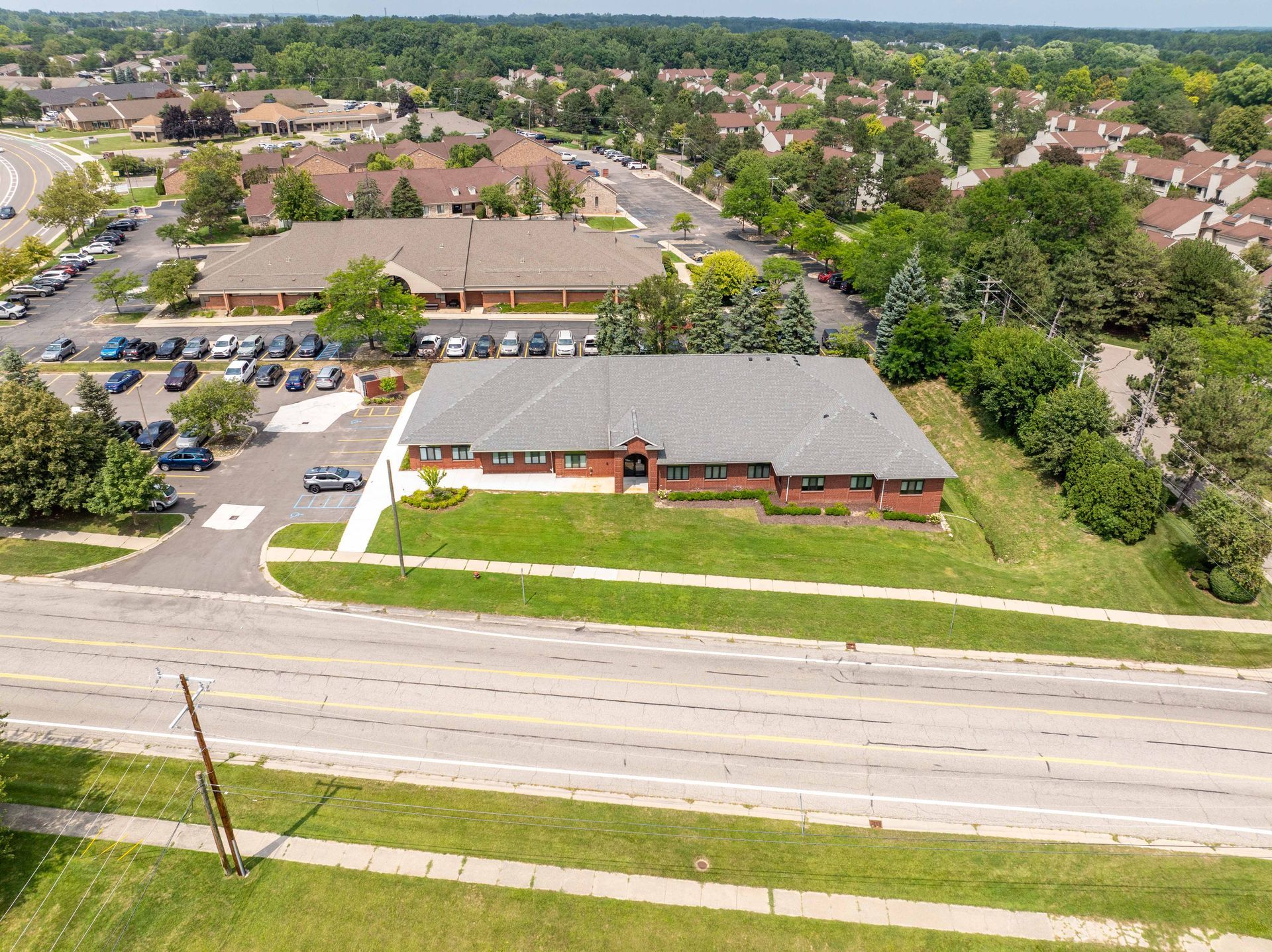 Aerial view of a brick building with a parking lot, set along a road with homes and trees in the background.