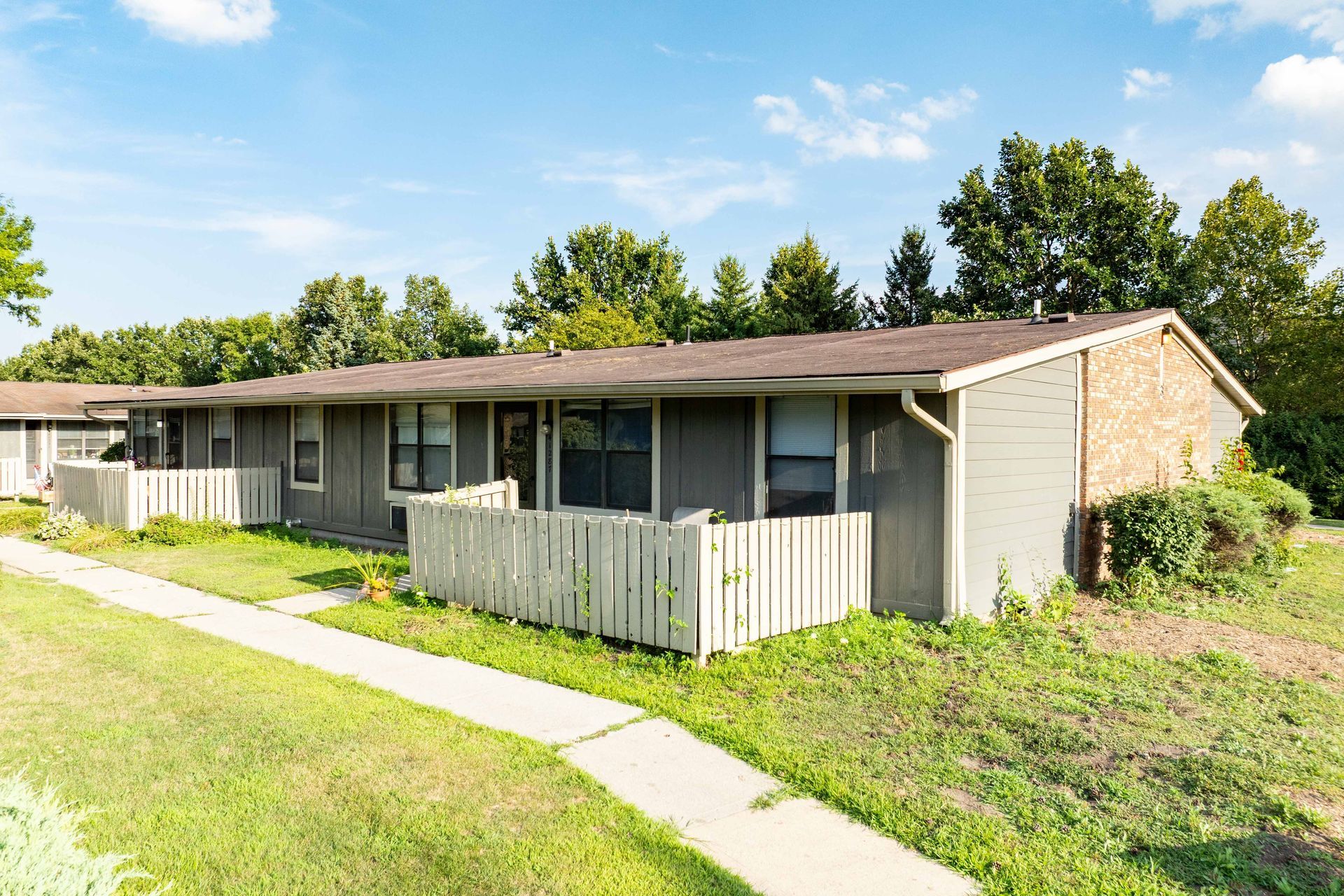 Gray and brick row houses with white fences and green grass under a blue sky.
