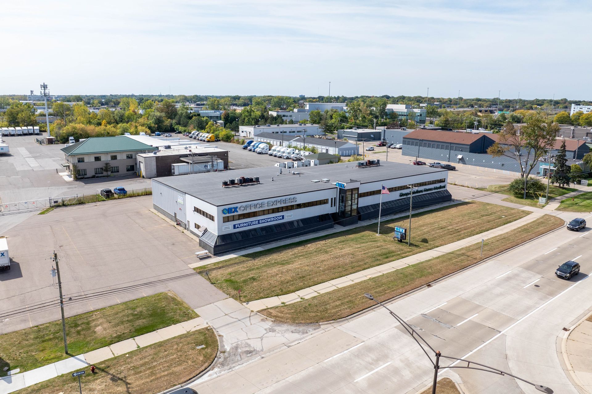 Aerial view of a two-story building with a gray roof, a large parking lot, and a green grass strip.