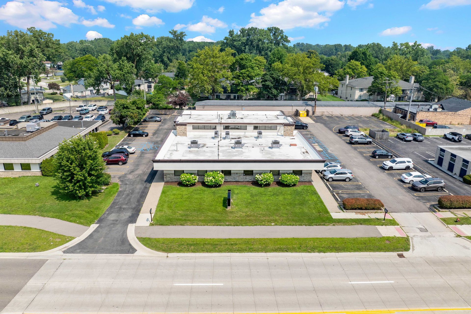 Aerial view of a low, white commercial building with cars parked around it, lush green lawn and trees.